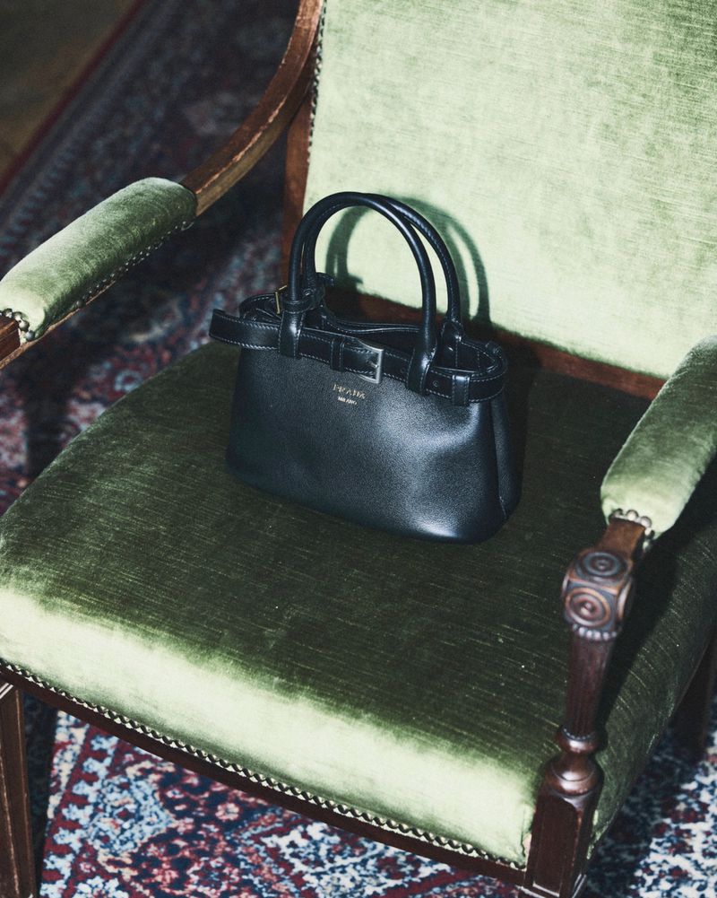 Black leather handbag resting on a vintage green velvet armchair with dark wooden trim