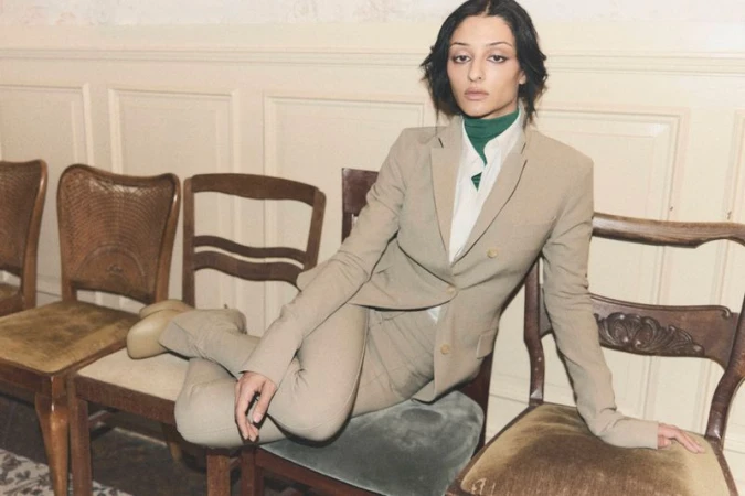 Sofie Royer seated cross-legged on wooden chairs wearing a beige suit and green turtleneck for Numéro Berlin by Emil Dietrich