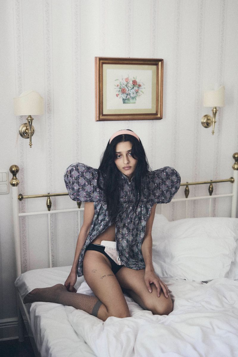 Woman kneeling on a white hotel bed wearing a patterned top with exaggerated puffy sleeves and a pink headband