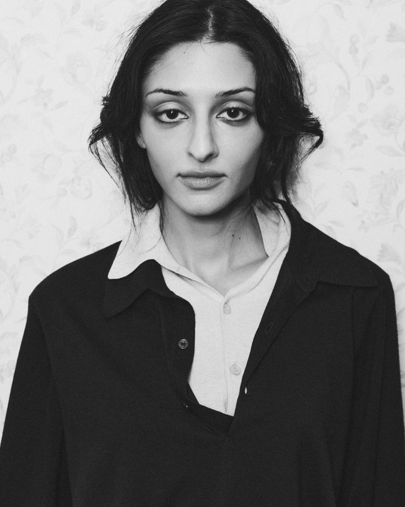 Black and white portrait of a woman with dark hair and heavy eye makeup wearing a dark buttoned shirt over a light collared top