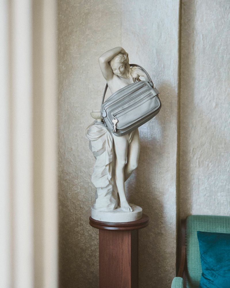 Classical marble statue on a wooden pedestal holding a grey leather handbag against a textured wall in a room.