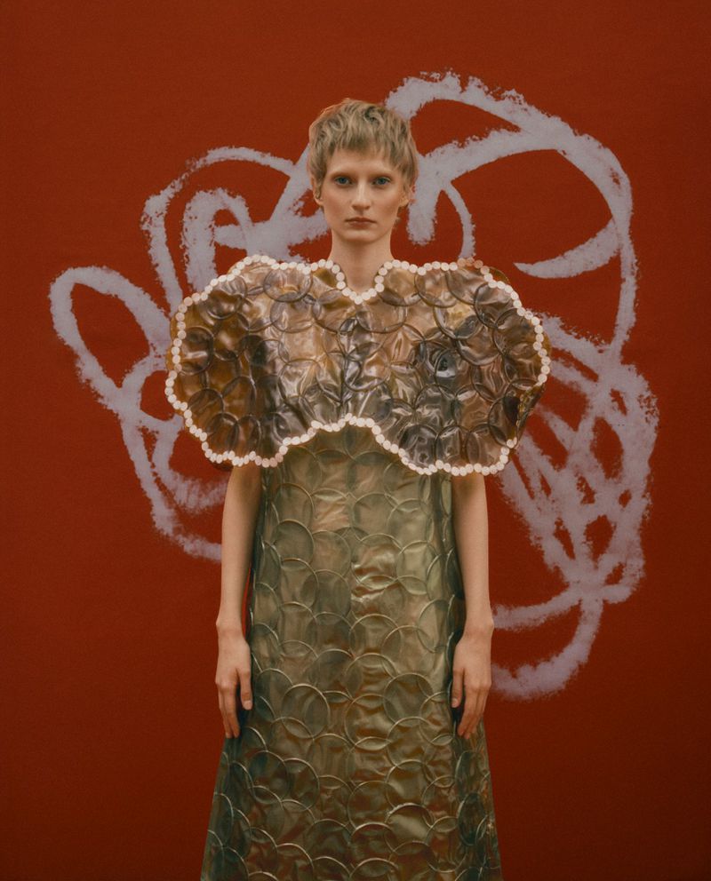 Model with short hair wearing a translucent bubble-textured dress against a dark red backdrop with pale pink painted scribbles.