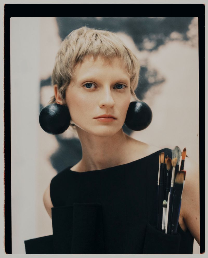 Portrait of a blonde woman wearing large round black earrings and a black top holding paintbrushes for Kinfolk