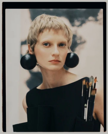 Portrait of a blonde woman wearing large round black earrings and a black top holding paintbrushes for Kinfolk