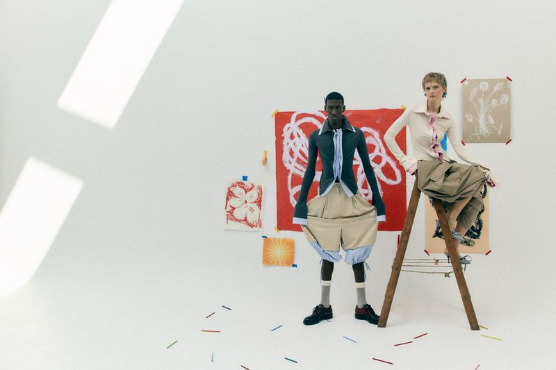 Two fashion models posing in a white studio with colorful artworks taped to the wall and crayons scattered on the floor.