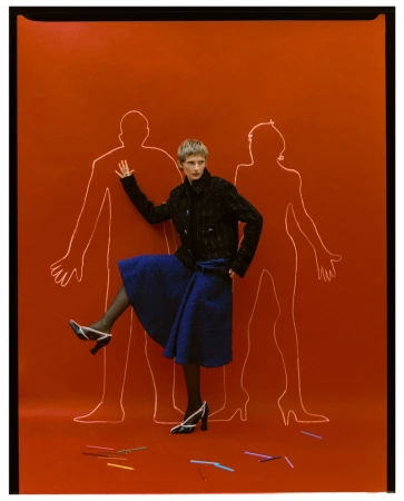 Model Anna Pavlova in black jacket and blue skirt posing before a burnt orange backdrop with chalk figure outlines.