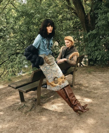 Two female models posing on a wooden park bench outdoors amidst lush green trees and foliage.