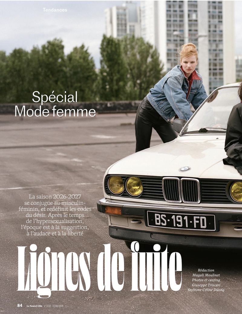 Model leaning on the hood of a vintage car in an outdoor parking lot setting with apartment buildings visible in the background