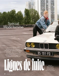 Model leaning on the hood of a vintage car in an outdoor parking lot setting with apartment buildings visible in the background