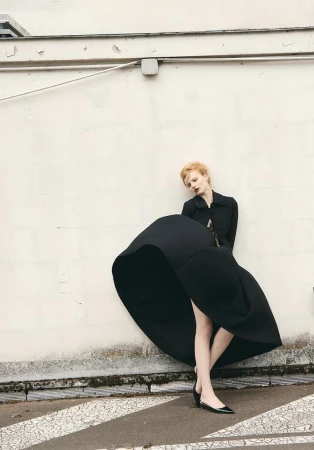 Model wearing a voluminous black skirt and matching jacket leaning against an off-white wall for L'Obs Magazine editorial.