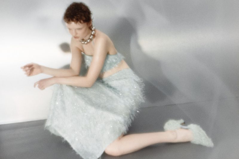 Model in pale blue textured two-piece outfit and chunky silver necklace kneeling against a grey backdrop for Tidal Magazine.