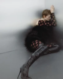 Model seated in the corner of a metallic grey room wearing a patterned long sleeve top and black thigh high boots