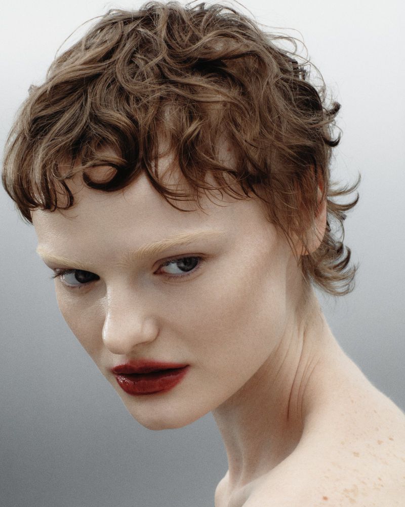 Close-up portrait of a model with short curly brown hair, dark lips, and a light background for Tidal Magazine.