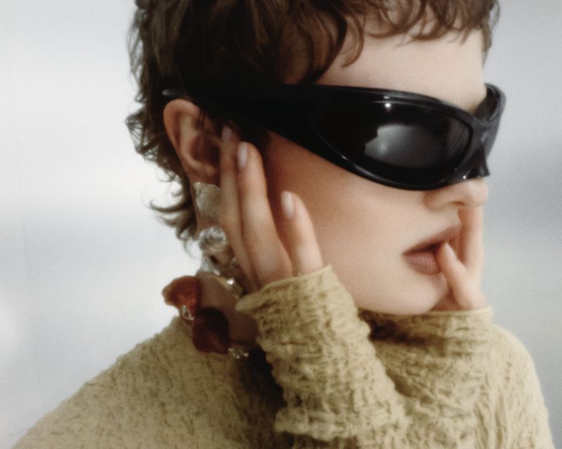 Model wearing dark wrap sunglasses and textured yellow top with chunky necklace against light background