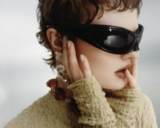 Model wearing dark wrap sunglasses and textured yellow top with chunky necklace against light background