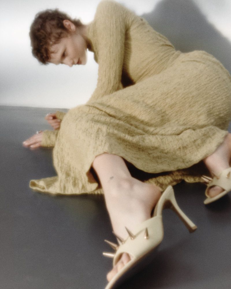 Model wearing a textured olive long-sleeved dress and heeled mules while seated against a grey backdrop for Tidal Magazine.