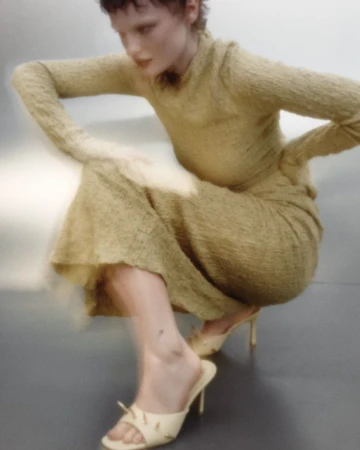 Model in textured olive green long-sleeve top and skirt with cream heeled mules squatting in a studio