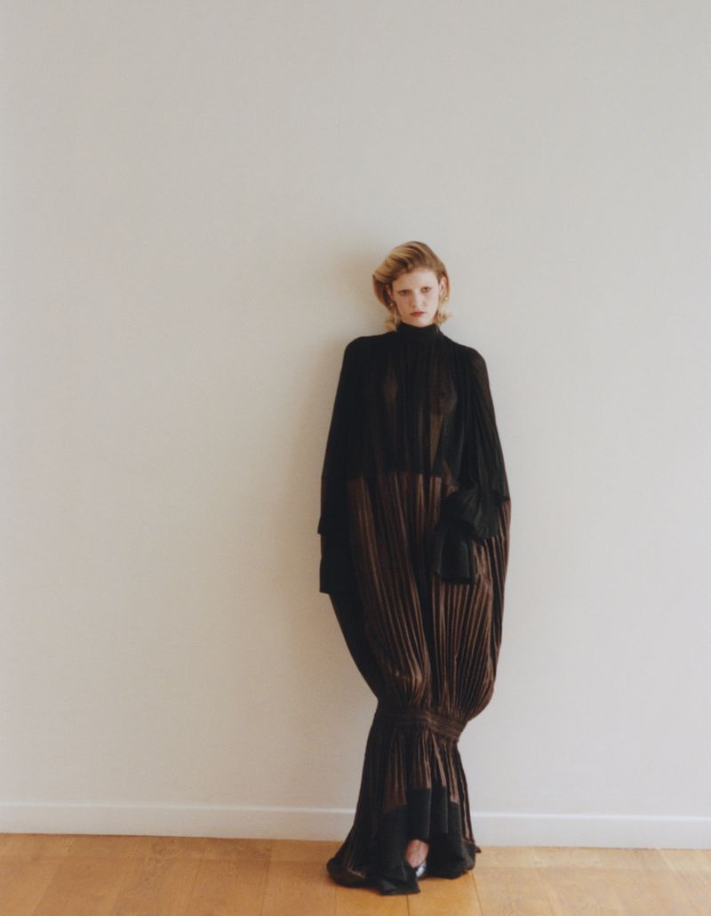 Model Camille Chifflot wearing a long black pleated gown standing against a neutral wall for 10 Magazine USA