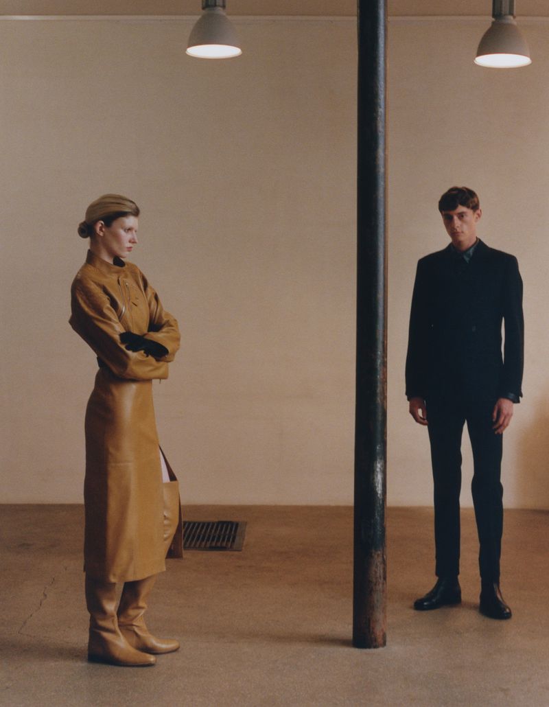 Woman in brown coat and boots standing beside a dark suit man in an interior space