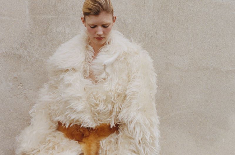Model wearing a voluminous cream shearling coat with a brown belt against a textured stone wall background.