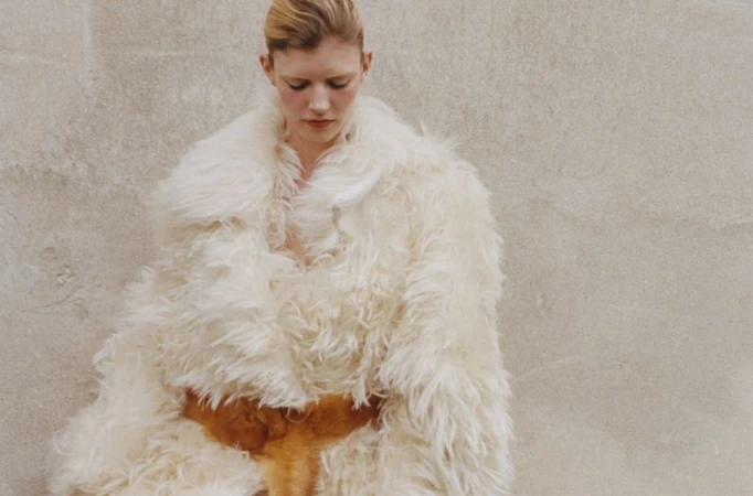 Model wearing a voluminous cream shearling coat with a brown belt against a textured stone wall background.