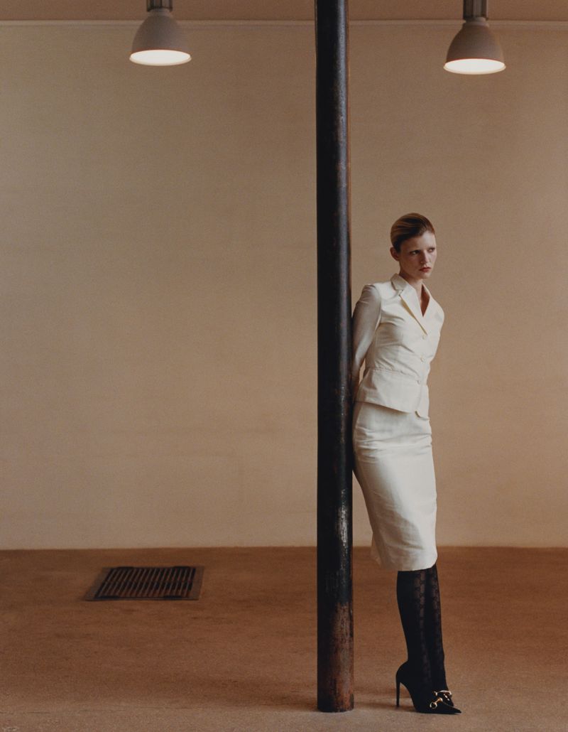 Model leaning against a dark vertical pillar in a neutral-toned room wearing a white skirt suit and black patterned tights