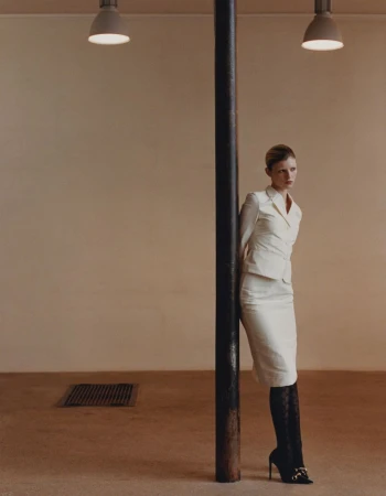 Model leaning against a dark vertical pillar in a neutral-toned room wearing a white skirt suit and black patterned tights