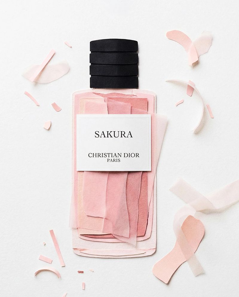 A stylized paper art Dior Sakura perfume bottle surrounded by pink paper cutouts on white background.