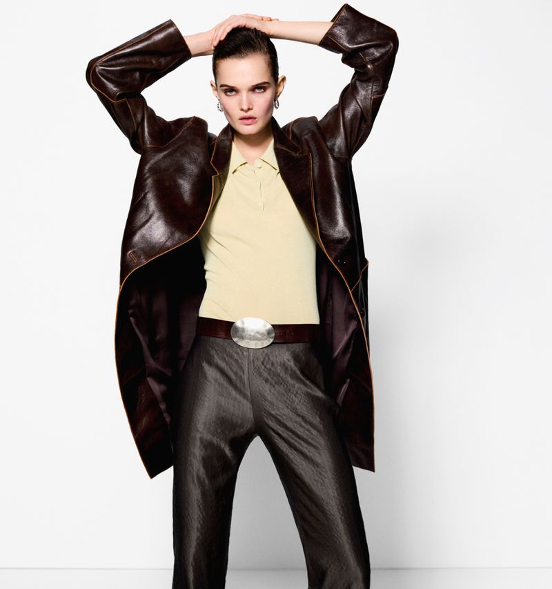 Adult woman poses in a studio wearing a brown leather jacket, light yellow polo shirt, dark brown pants, and a silver belt buckle.