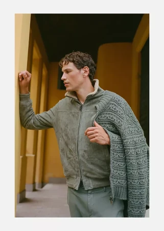 Model Paul Hupfer leaning against a yellow architectural column wearing a shearling-lined jacket and holding a knit sweater.