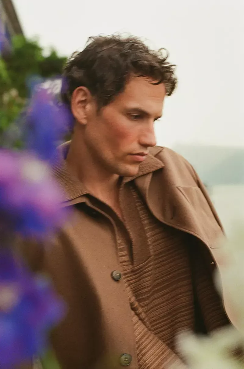 Model Paul Hupfer in a brown collared jacket and textured knit polo outdoors with blurred blue flowers in the foreground.