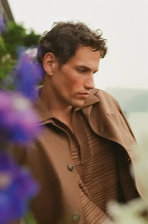 Model Paul Hupfer in a brown collared jacket and textured knit polo outdoors with blurred blue flowers in the foreground.