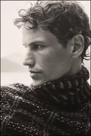 Portrait of model Paul Hupfer wearing a textured dark turtleneck sweater in profile view against an overcast background