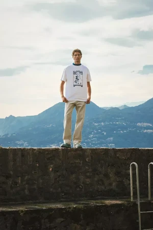 Model Paul Hupfer standing on a stone ledge outdoors wearing a white graphic t shirt and light trousers against mountain and lake view