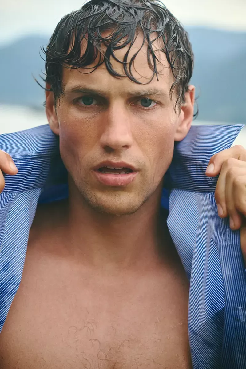 Close-up portrait of model Paul Hupfer with wet dark hair holding up the collar of a striped blue shirt.