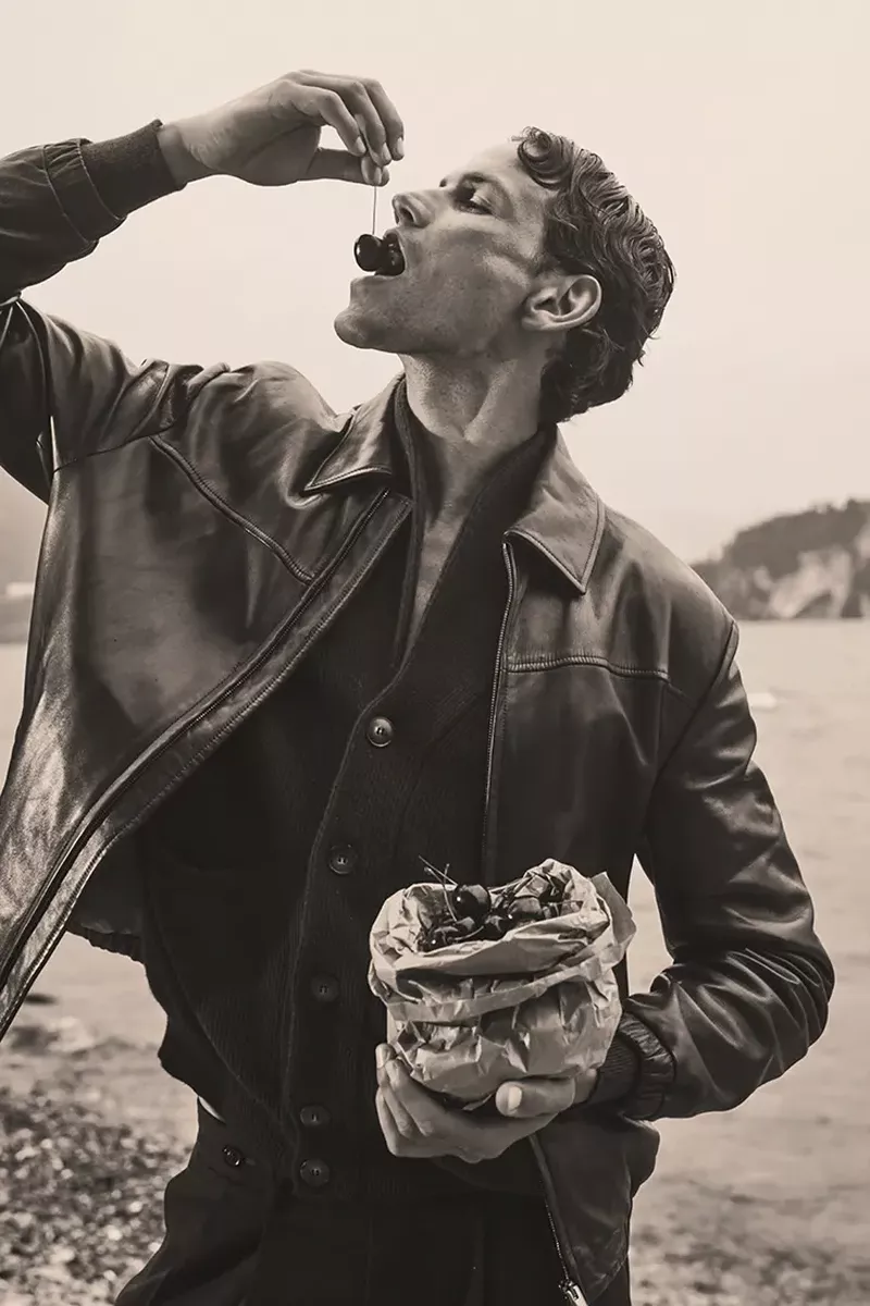 Model Paul Hupfer in a leather jacket eating a cherry outdoors by a lake for MMScene in The Last Weekend editorial.
