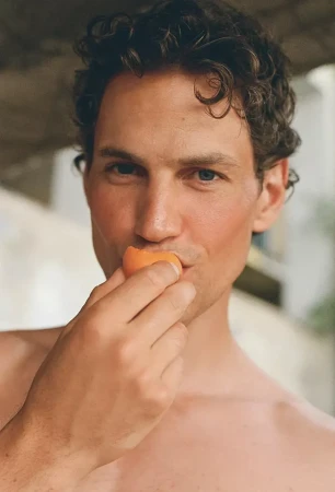 Close-up portrait of model Paul Hupfer holding a piece of fruit to his lips against a neutral background.