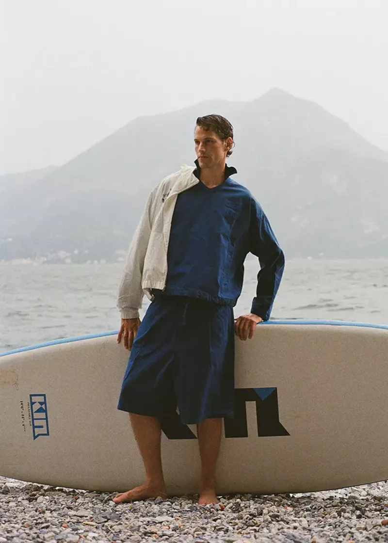Model Paul Hupfer standing on a pebble shore holding a surfboard with a misty mountain and water background.