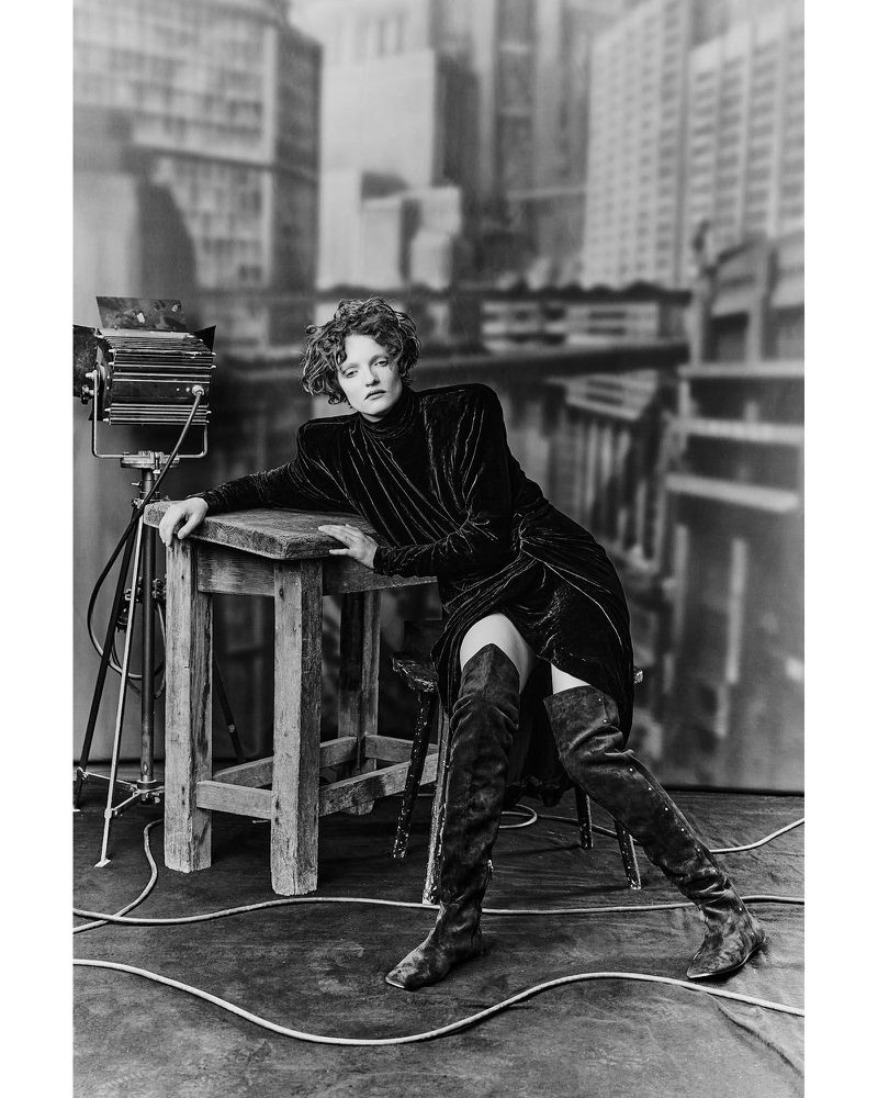 Model in black dress and tall boots leaning on a wooden table in a studio with a city backdrop.
