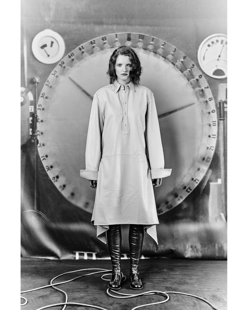 Monochrome portrait of a model wearing a collared dress and boots against a large clock-face backdrop