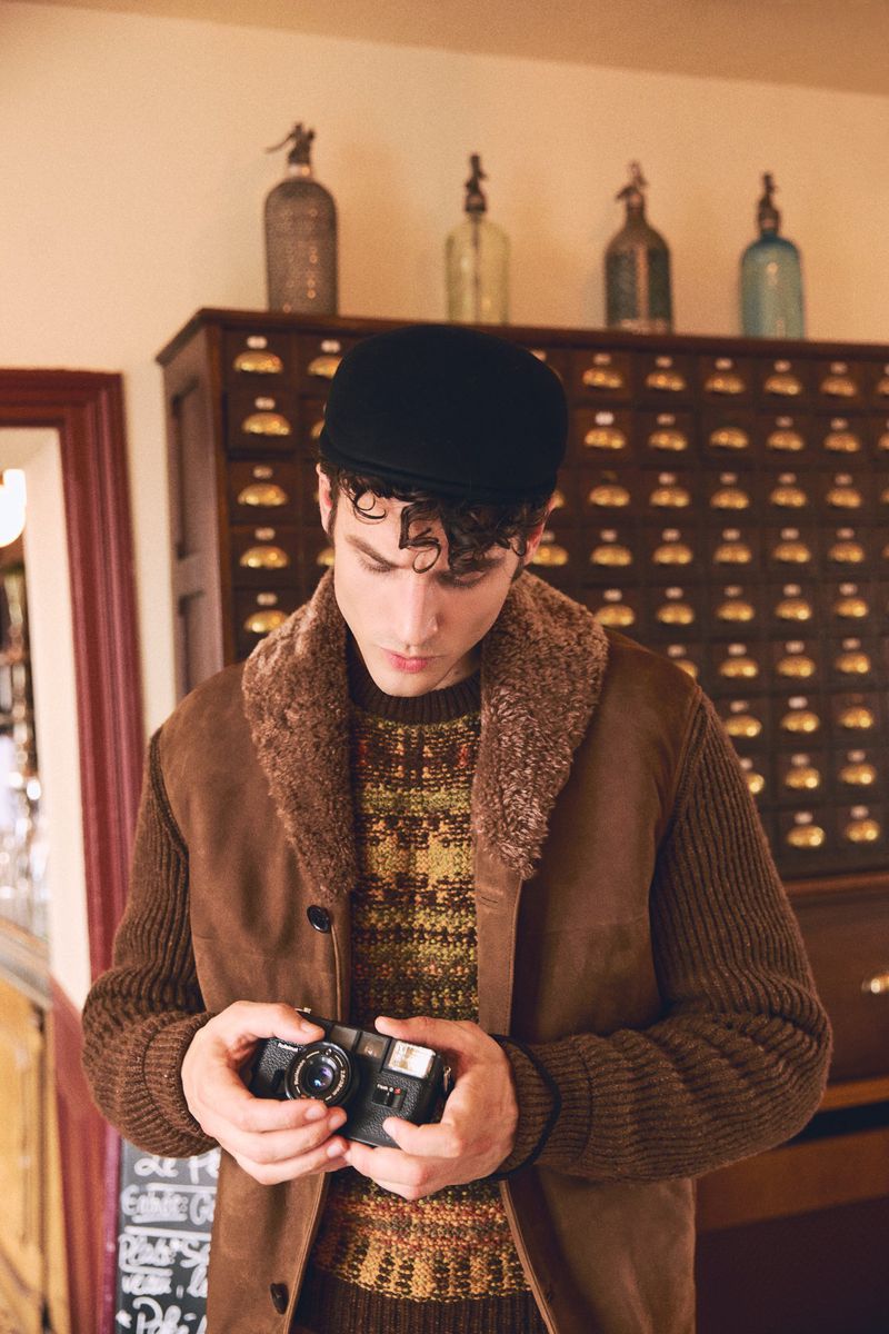 Man wearing a brown shearling collar jacket and black flat cap holding a vintage camera in an interior setting