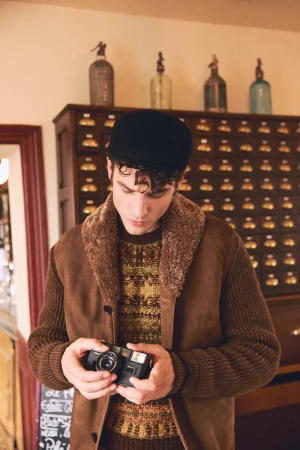 Man wearing a brown shearling collar jacket and black flat cap holding a vintage camera in an interior setting