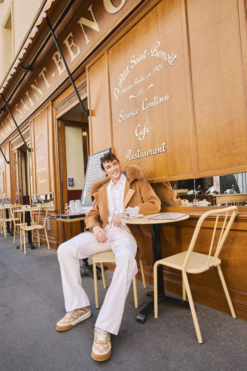 Model Federico Novello seated outside a cafe wearing a camel coat and white trousers for Marie Claire Czech Republic.