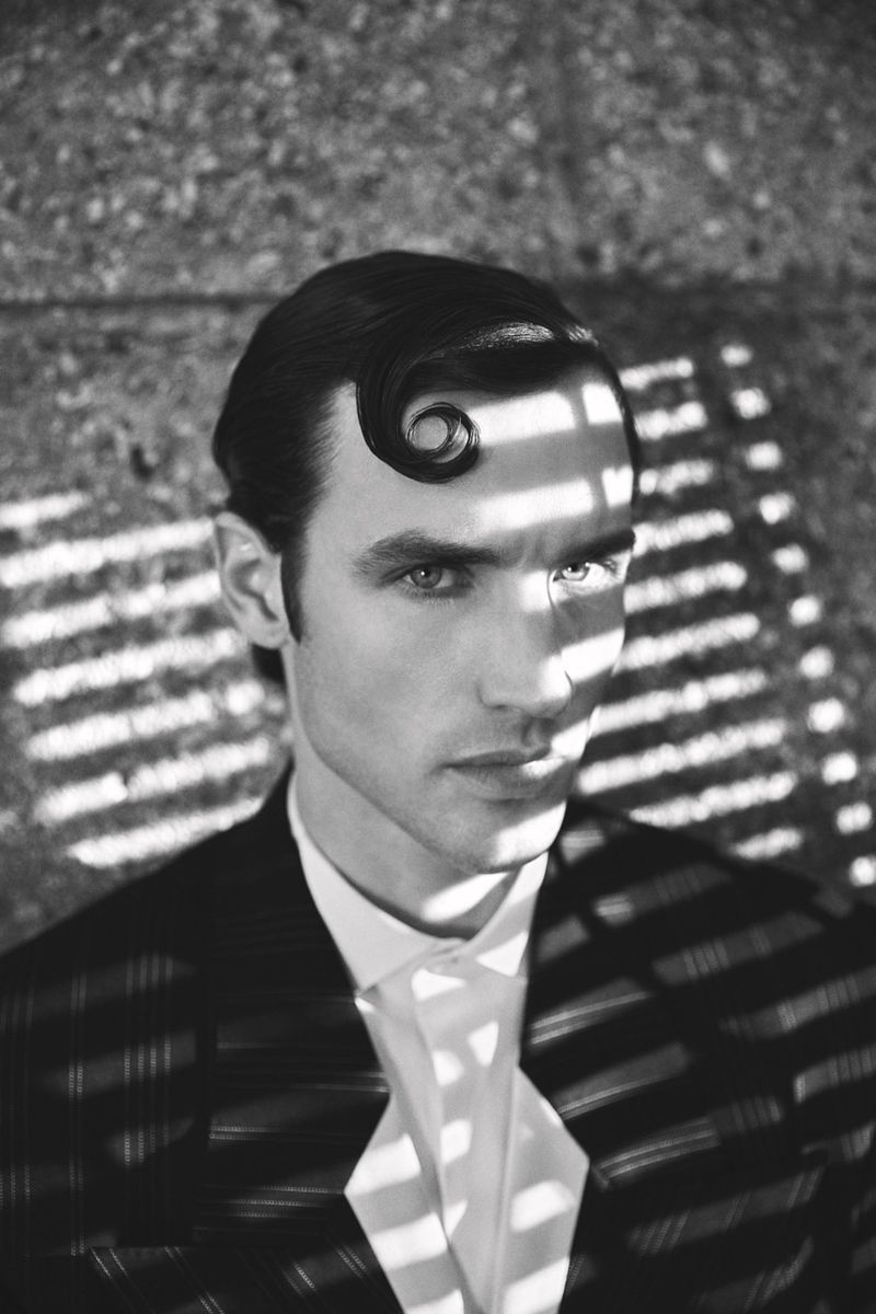 Black and white portrait of a man with a sculpted curl on his forehead, wearing a patterned jacket and white shirt.