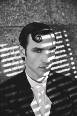 Black and white portrait of a man with a sculpted curl on his forehead, wearing a patterned jacket and white shirt.
