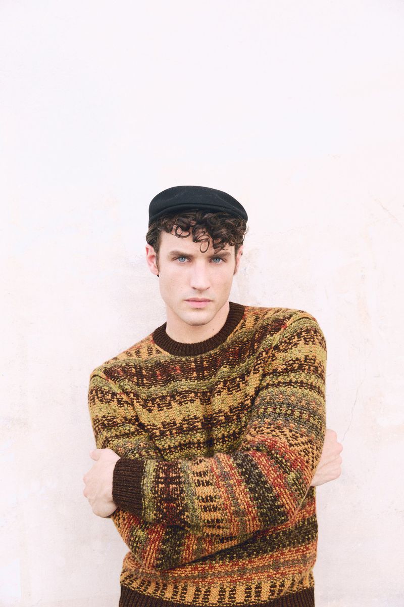 Model wearing a patterned knit sweater and dark flat cap with arms crossed against a plain white wall
