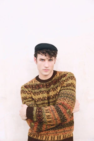 Model wearing a patterned knit sweater and dark flat cap with arms crossed against a plain white wall