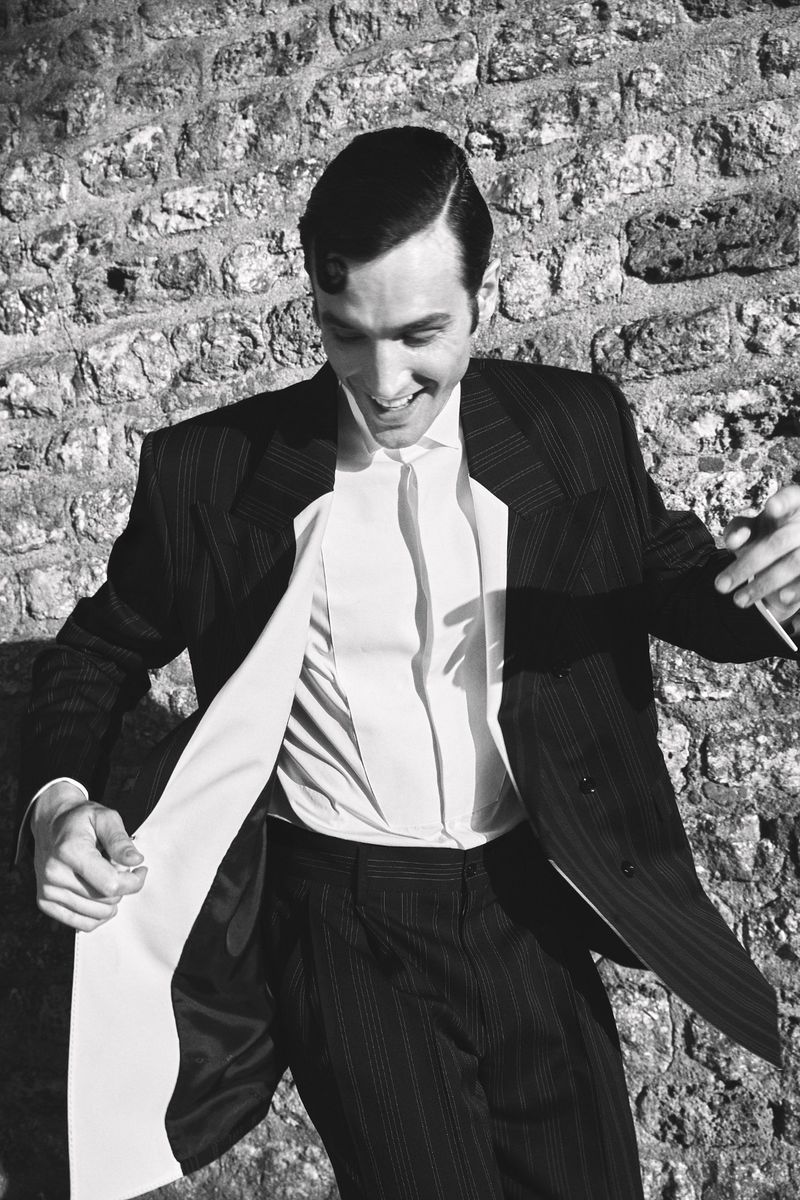 Monochrome portrait of a smiling man wearing a dark pinstripe suit and white shirt against a textured stone wall