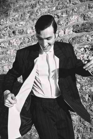 Monochrome portrait of a smiling man wearing a dark pinstripe suit and white shirt against a textured stone wall