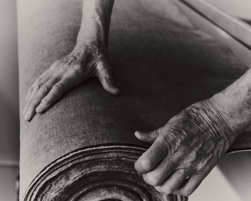 Close-up monochrome image of elderly hands unrolling a thick roll of textured fabric material.